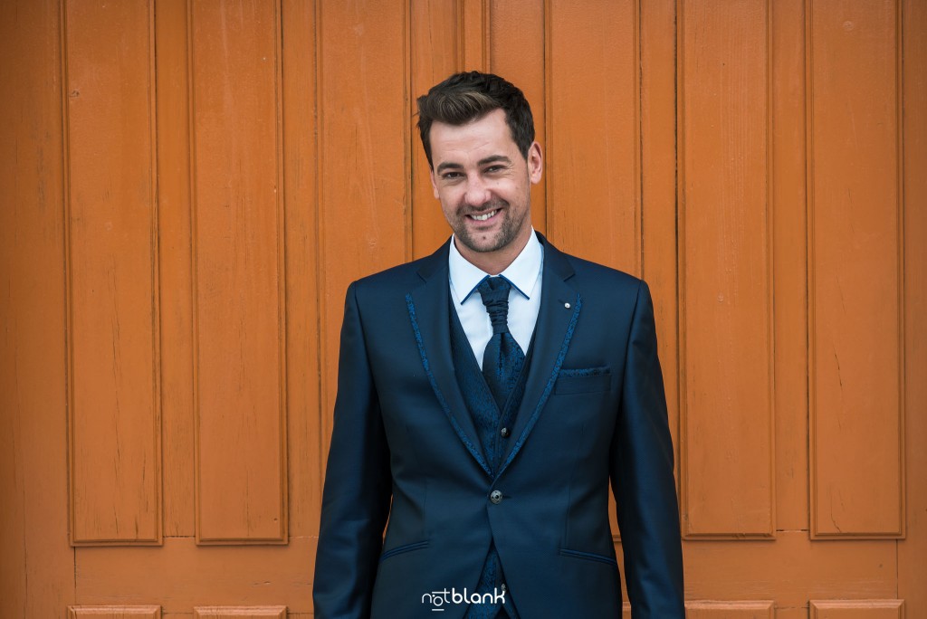 Boda en Salceda de Caselas. Retrato del novio vestido con su traje de boda azul con una puerta de madera al fondo. Reportaje realizado por Notblank fotógrafos de boda en Galicia.